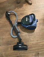Image showing a blue canister vacuum with attached hose, wand, and floor cleaning head resting on a wooden floor.