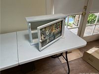 White wooden curio shelf with a glass front panel, photographed open on a white folding table next to a door with glass panes.