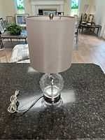 Photo of a desk lamp with clear glass bubble textured base and beige fabric shade on a dark granite countertop inside a bright living room.