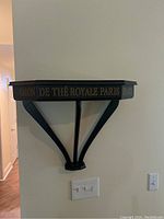 Wall-mounted black wooden shelf with gold text reading 'Salon De Thé Royale Paris'. Curved support brackets visible.