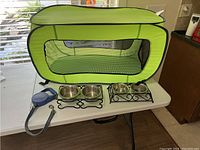 Green collapsible pet carrier, two pairs of stainless steel feeding bowls in black holders, and blue retractable leash on white table.