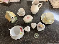 Various small collectibles on a dark surface including porcelain cups and saucers, a pitcher, heart-shaped trinket boxes, a floral brooch, and a small metal card holder.