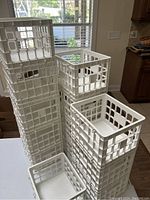Six white plastic mini milk crate style storage containers of varying sizes stacked in a kitchen setting near a window and countertop.