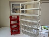 Photo showing a white plastic mini shelving unit with five shelves and a red plastic stacked basket storage unit with six compartments, placed indoors next to a framed picture on a wall.