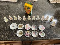 Photo showing 12 miniature coffee cups with saucers in a row, each cup having a different colorful design. Also displayed are 10 small decorative plates with various paintings. Crayola crayon box placed for size comparison, indicating the very small scale of the items.