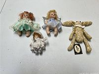 Four handheld size dolls laid out on white surface: three porcelain dolls with fabric clothing and one plush dog doll with collar and tag.