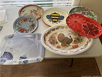 View of seven various decorative platters and plates arranged on a table showing different colorful designs including a turkey, flowers, bee, fruits, beach theme, and fall foliage.