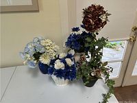 Photo of five silk flower arrangements in various vases including blue, white, and maroon flowers with greenery displayed on a white table.