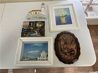 Top view of a white table showing five wall art pieces arranged closely: three framed pictures, one decorative plaque with deer, and one 3D lighthouse sculpture.