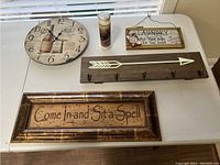 Display of five wall decor items on a white table including a clock, candle, two plaques, and a wooden hook rack with an arrow.