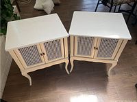 Two white wooden nightstands side by side, showing tops and front cabinets with woven cane doors and curved legs on a brown wood floor.