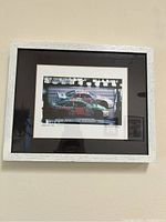 Framed print showing a race car with a black mat and white textured frame hung on a wall.