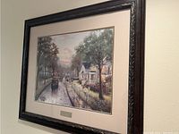 Photo of a framed Thomas Kinkade library edition picture, showing a street scene with houses, trees, and people. The frame is dark wood with ornate carvings and measures about 26 by 30 inches.