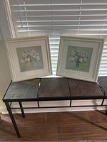 Two framed floral pictures, each 16x16 inches, resting on a metal and stone bench by window blinds.