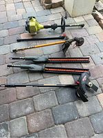 Photo showing Ryobi weed eater powerhead with multiple attachments including two hedge clippers, a yellow hedge clipper, a Ryobi head attachment, and an extension pole placed on paved ground.