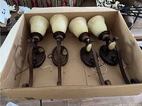 Four large and heavy wall sconces arranged in a cardboard box, showing dark metal mounting plates and curved arms with exposed wiring and cream colored bell-shaped glass shades.