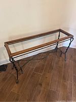 Photo of wooden and metal sofa or console table frame missing glass top, placed on a wooden tiled floor against a beige wall.