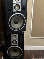 Pair of vintage Sanyo stereo speakers stacked vertically against a wall. Each speaker has a large woofer at the bottom, midrange drivers, and circular sound ports. Black rectangular cabinets approximately 30 inches tall.