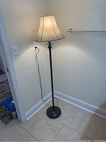 Full view of the vintage freestanding floor lamp standing near the corner of a tiled floor room. It has a cream-colored fabric lampshade and dark metal pole and base. Plugged into electrical outlet for power.
