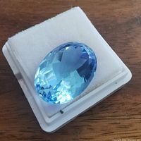 Close-up of the oval, faceted blue quartz gemstone with bright clarity on a white cushioned holder, showing the stone's cut and color.