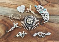 Eight costume brooches displayed on a wooden surface showing various styles including heart-shaped and dancer designs with rhinestones and colored stones.