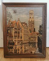 Front view of framed vintage needlepoint artwork showing detailed architectural scene with buildings and clock tower reflecting in water.