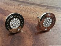 Pair of round gold tone Michael Kors stud earrings with clear crystals and logo engraving on a wooden surface.