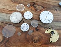 Overview of pocket watch parts on wooden surface showing dials, crystals, and movement plate with components.