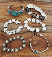 Top-down photo showing six assorted bracelets displayed on a wooden surface. Includes turquoise floral cuff and various silver-tone and bronze-tone bracelets.