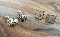 Two pairs of cufflinks on a wooden surface; one pair square with opaline stones, the other round with black and gold floral design.
