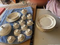 Top view of all china pieces laid out on a blue cloth showing cups, sugar bowl with lid, lidded serving bowl, and stacked dinner plates with floral patterns and gold trim.