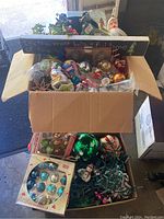 Open box filled with assorted vintage Christmas decorations, ornaments in varied colors, and a visible metal Christmas tree stand behind the box.