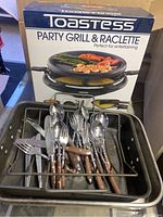 Toastess Party Grill and Raclette cooker with box, set of silverware with wooden handles, and 2 metal roasting pans seen from front.