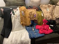 Image showing various sizes of new clothing items with tags laid out on a couch, including dresses, skirts, tops, and pants in colors like black, white, mustard yellow, denim blue, pink, and olive green.