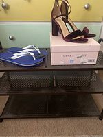 Black metal shoe rack with perforated shelves, a pair of burgundy Ivanka Trump high heels on the top shelf with the original box, and a pair of blue Havaianas flip flops alongside.