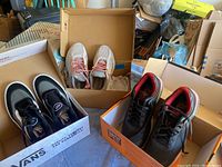 Image shows three pairs of shoes in their boxes placed on a blanket; black Vans sneakers on the left, grey/pink athletic shoes in the center box, and black/red sneakers on the right.