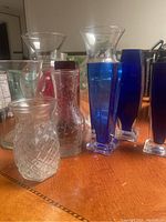 Full view of the assortment of glass vases on a wood table showing various shapes, sizes, and colors including clear and blue glass pieces.