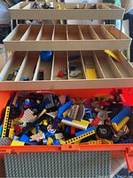 Photo showing the large orange tackle box open with the bottom compartment filled with assorted Lego bricks and pieces in multiple colors and shapes, along with beige plastic sorting trays above with some small items inside.