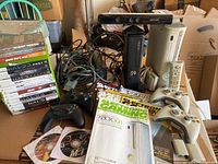Photo of two Xbox 360 consoles, Xbox Kinect, multiple controllers, cables, magazines, and a large stack of games.