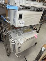 Two Kenmore air conditioning units stacked on metal shelving rack. One unit on top, one below. Both white with digital controls on front panel left side. Control buttons include power, timer and mode options.