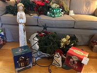 Overview photo showing tall stone angel statue, boxed porcelain lighted house, metal tree stand with greenery and ribbon, red poinsettias behind, and assorted garland pieces.
