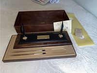 Full desk pen set displayed in original box including two pens, leather and wood base with brass plate, two pen holders, paperwork and spare cartridge.