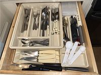 Drawer with organized stainless steel flatware and utensils, including forks, spoons, knives with white and black handles, plastic measuring spoons, and cutting utensils.