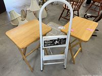 Shows two folding wooden side tables with natural wood tops and cross support legs, white metal folding step stool with handle, and white metal mesh square table