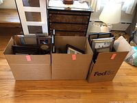 Three large cardboard boxes filled with assorted picture frames of different sizes and colors on a wooden floor.