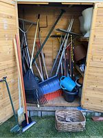 Wooden outdoor shed with various large garden tools including rakes, snow shovels, pitch fork, a watering can, black plastic pots, and a basket.