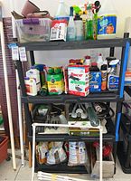 Three-shelf black plastic shelving unit holding various gardening products including bottles, boxes, bags, hoses, and pots.