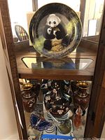 Three metal vases flanking a larger metal vase, two glass animal figurines (bird and cat), a blue butterfly figurine, and a panda collectable plate in the background.