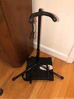 Black metal guitar stand with padded yoke and three legs, shown standing on wooden floor next to wall.