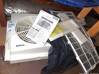 Holmes window fan with white body, power cord, filter unit and instruction manual placed on top. Styrofoam buffer under the fan.